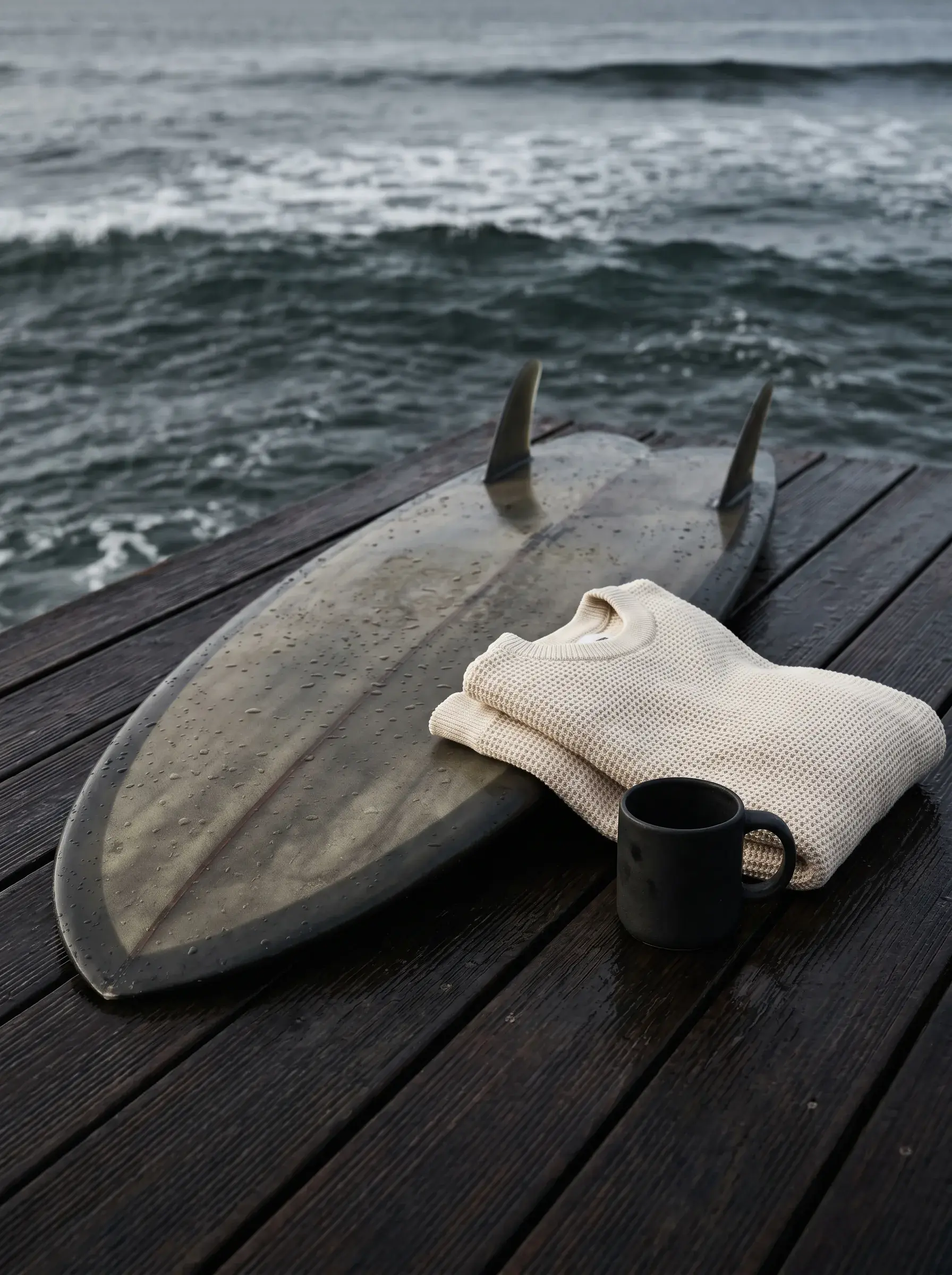 Planche de surf avec dérives, pull roulé et mug noir sur terrasse en bois face à l'océan
