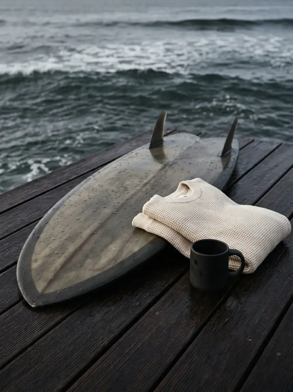 Planche de surf avec dérives, pull roulé et mug noir sur terrasse en bois face à l'océan