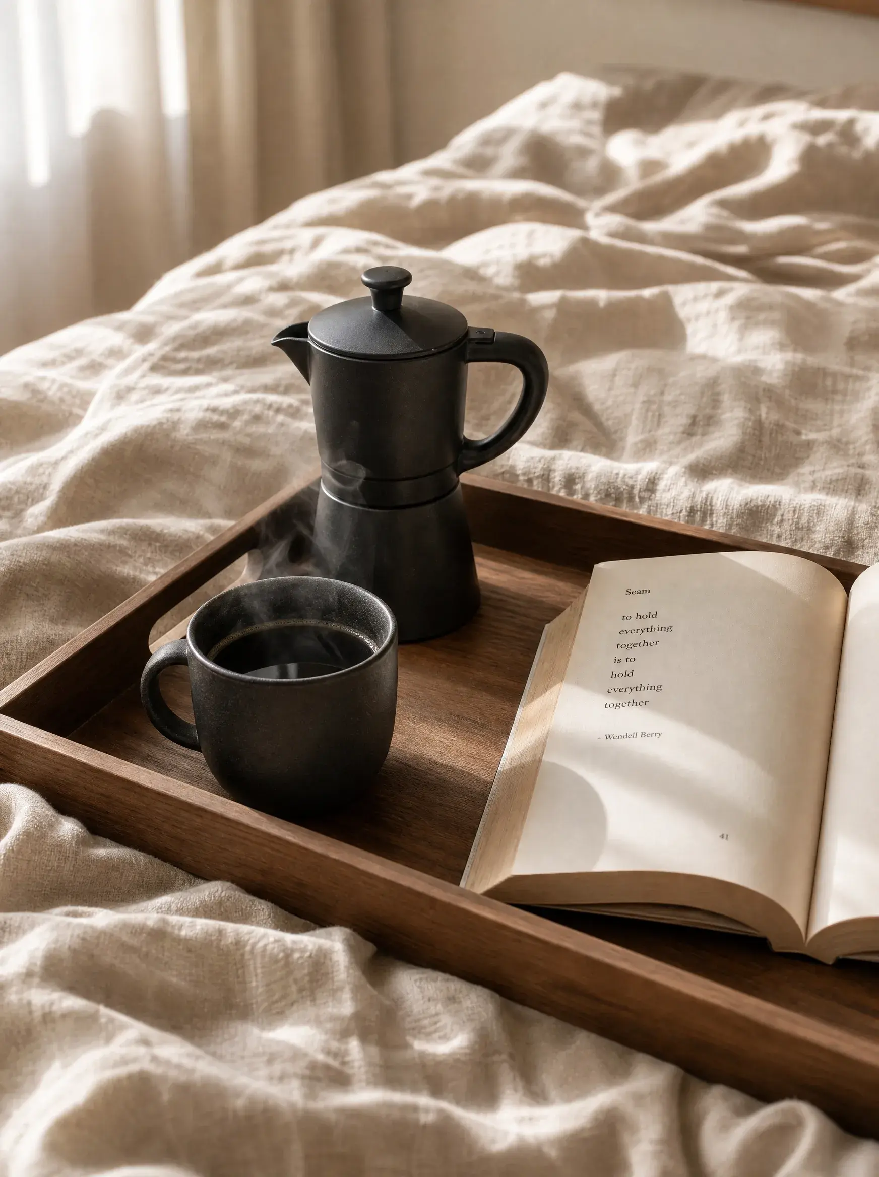 Plateau bois dans lit – cafetière moka noire, mug fumant, livre ouvert, linge beige froissé