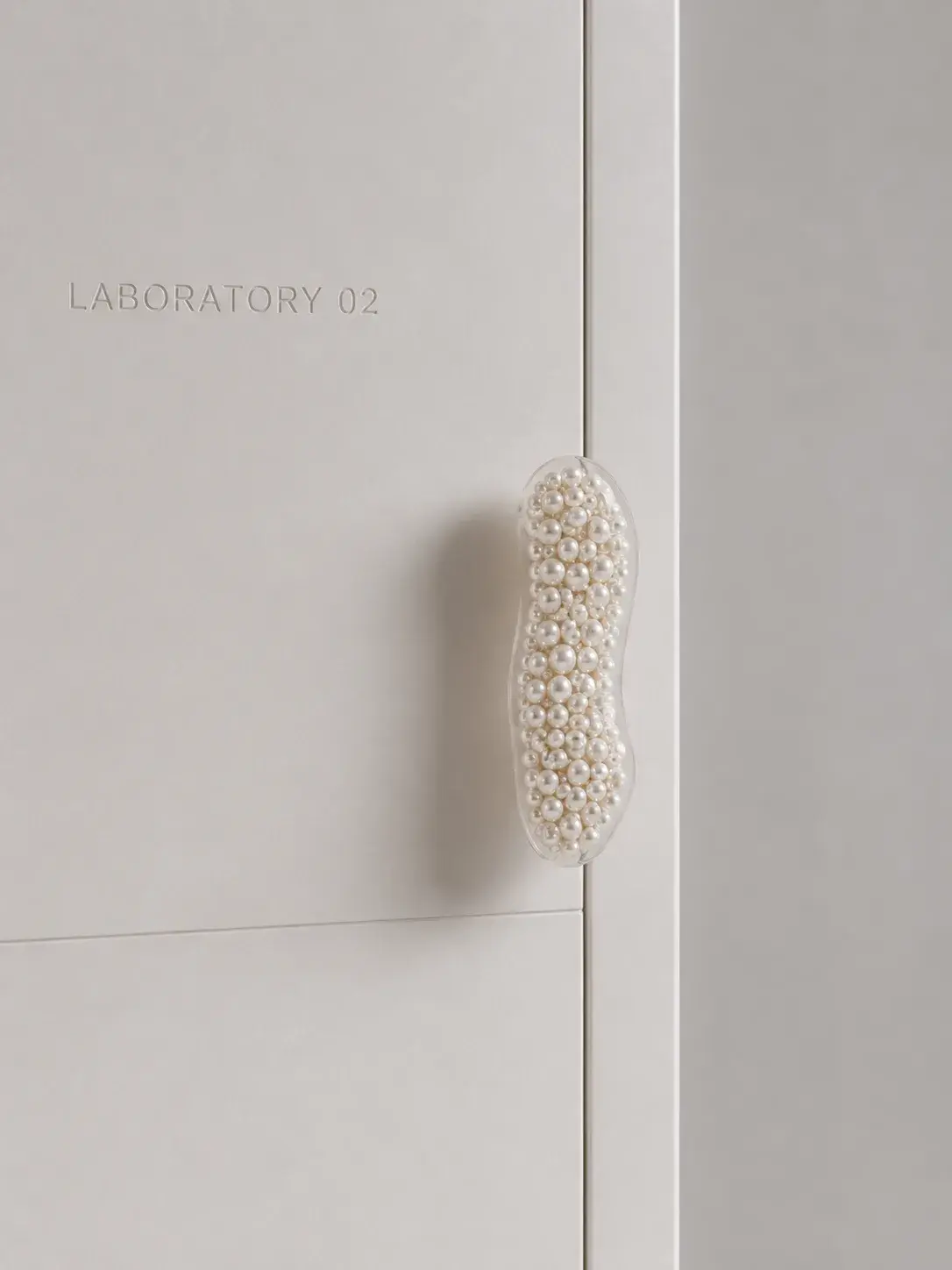 Poignée de porte recouverte de perles calibrées sur porte blanche marquée LABORATORY 02, détail nacré