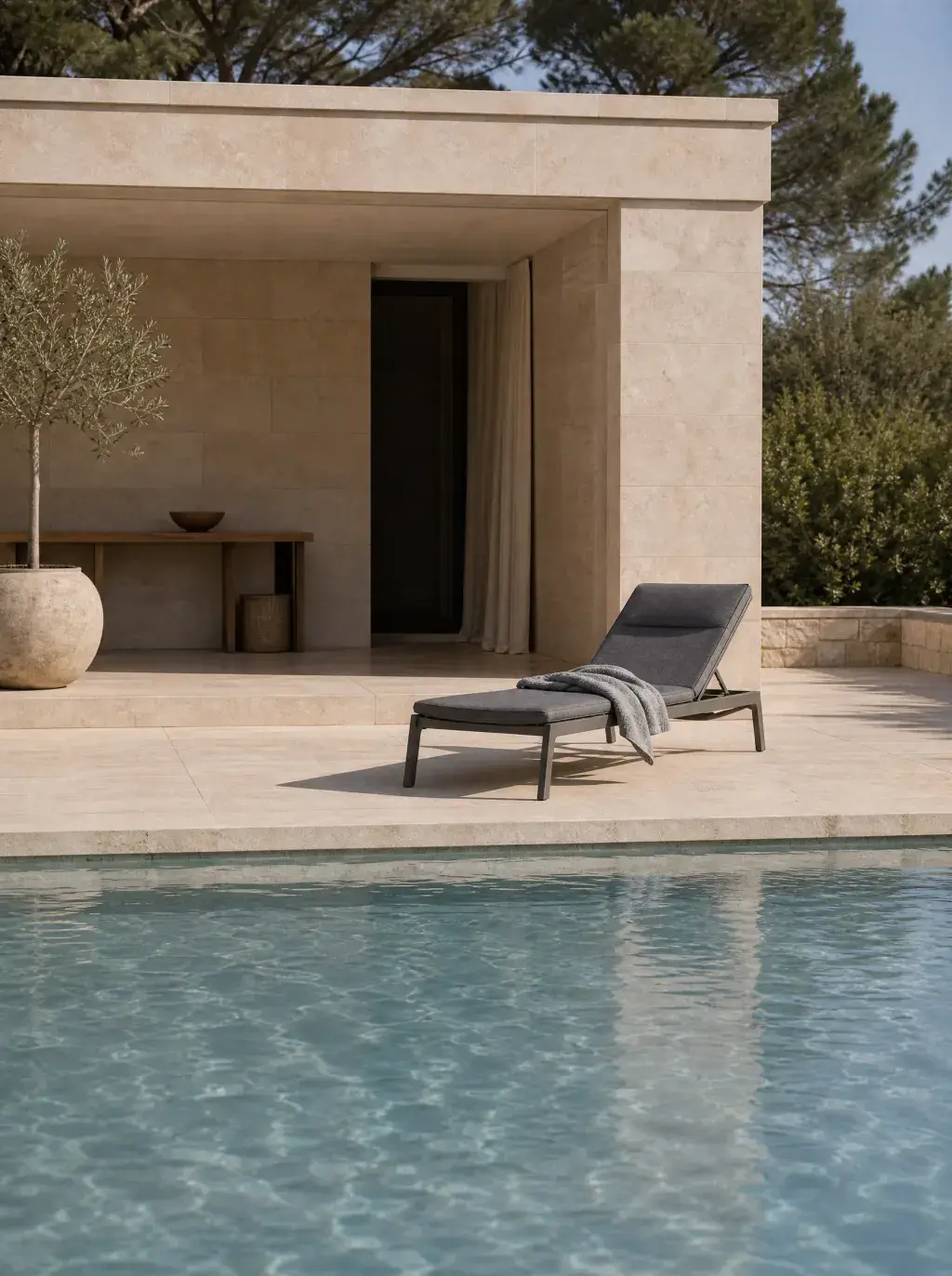 Pool house minimaliste en pierre calcaire avec piscine à débordement, chaise longue et olivier en Provence