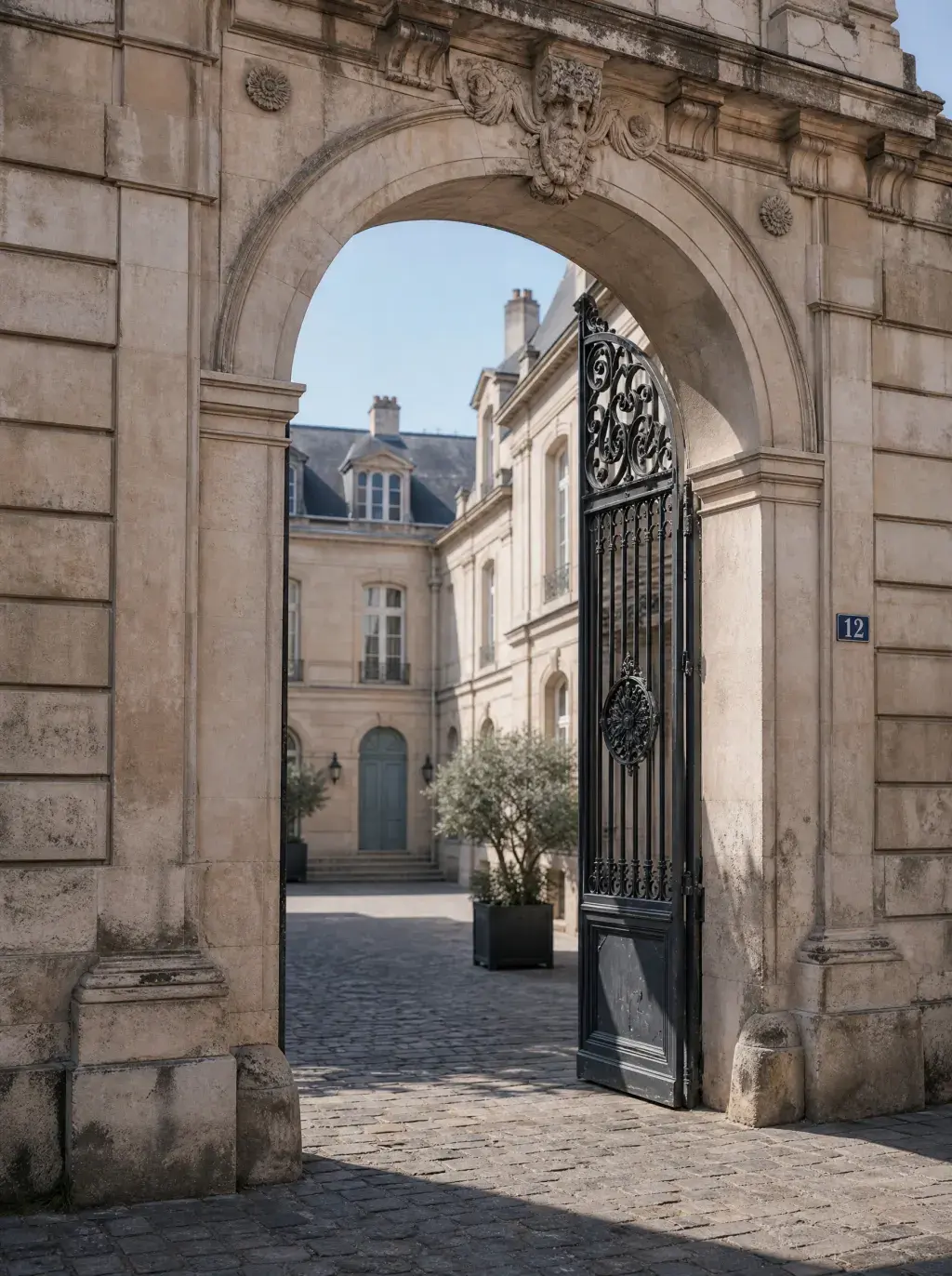 Portail en fer forgé d'un hôtel particulier parisien ouvert sur une cour pavée avec olivier et plaque de rue