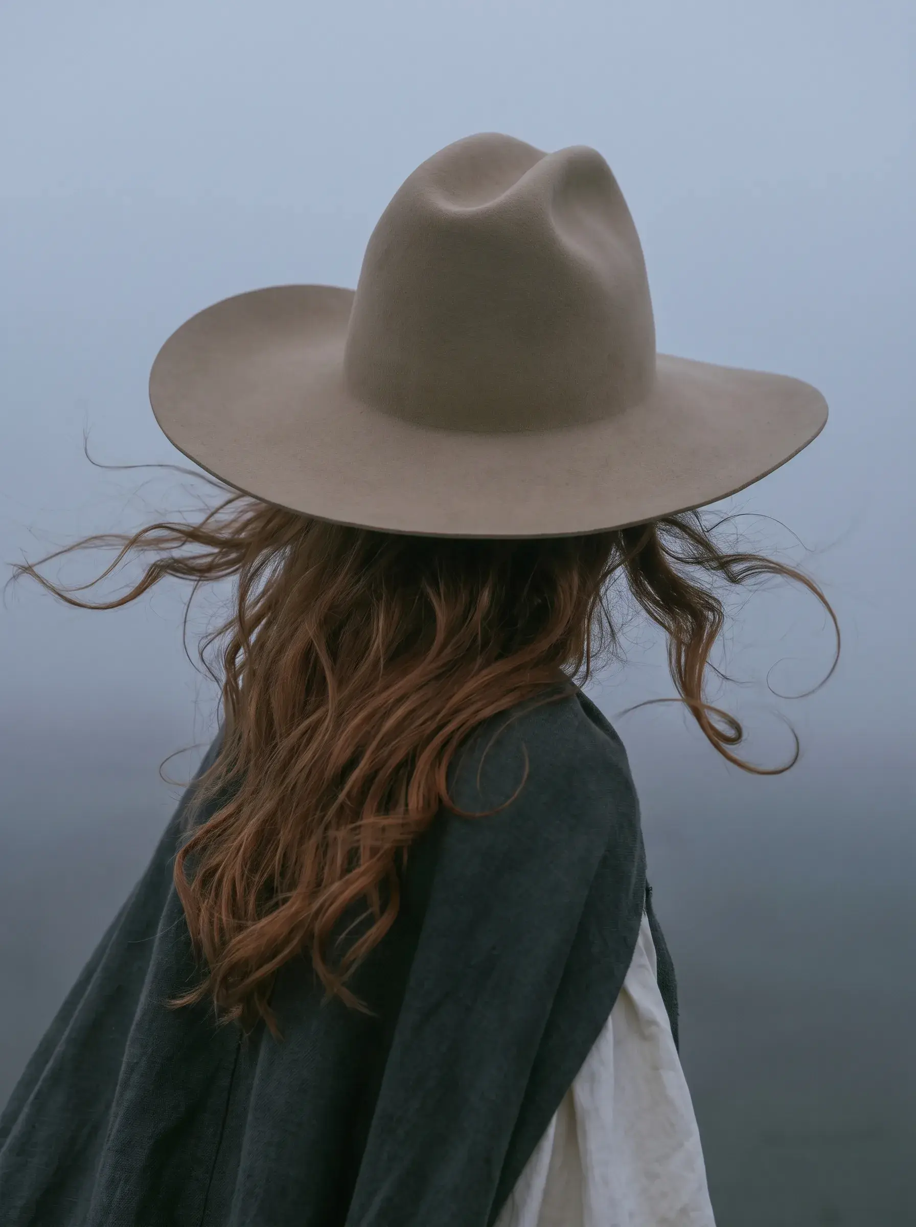 Portrait de dos d'une cavalière rousse en cape sombre et chapeau beige dans la brume grise