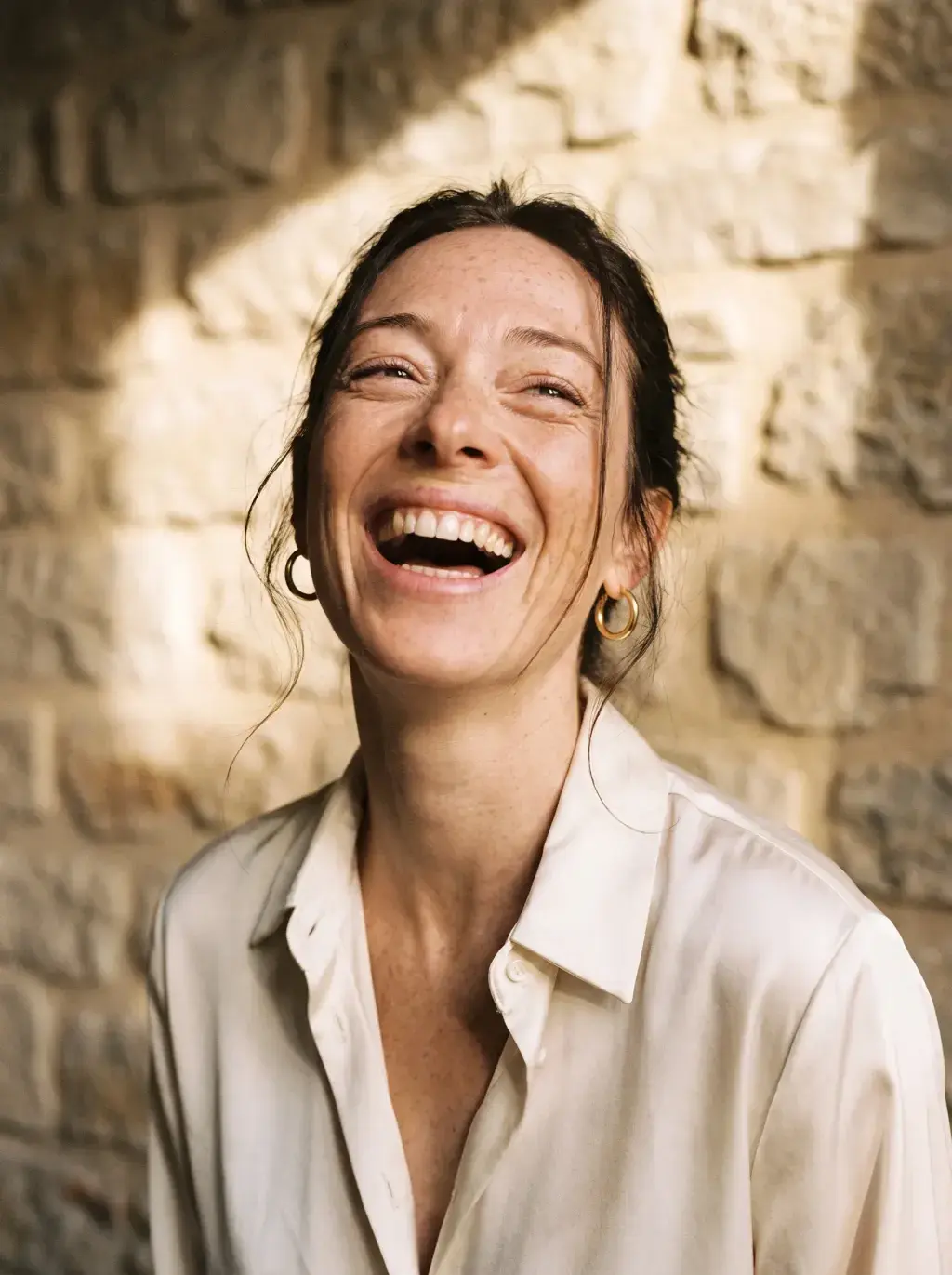 Portrait de femme brune riant aux éclats devant un mur en pierre
