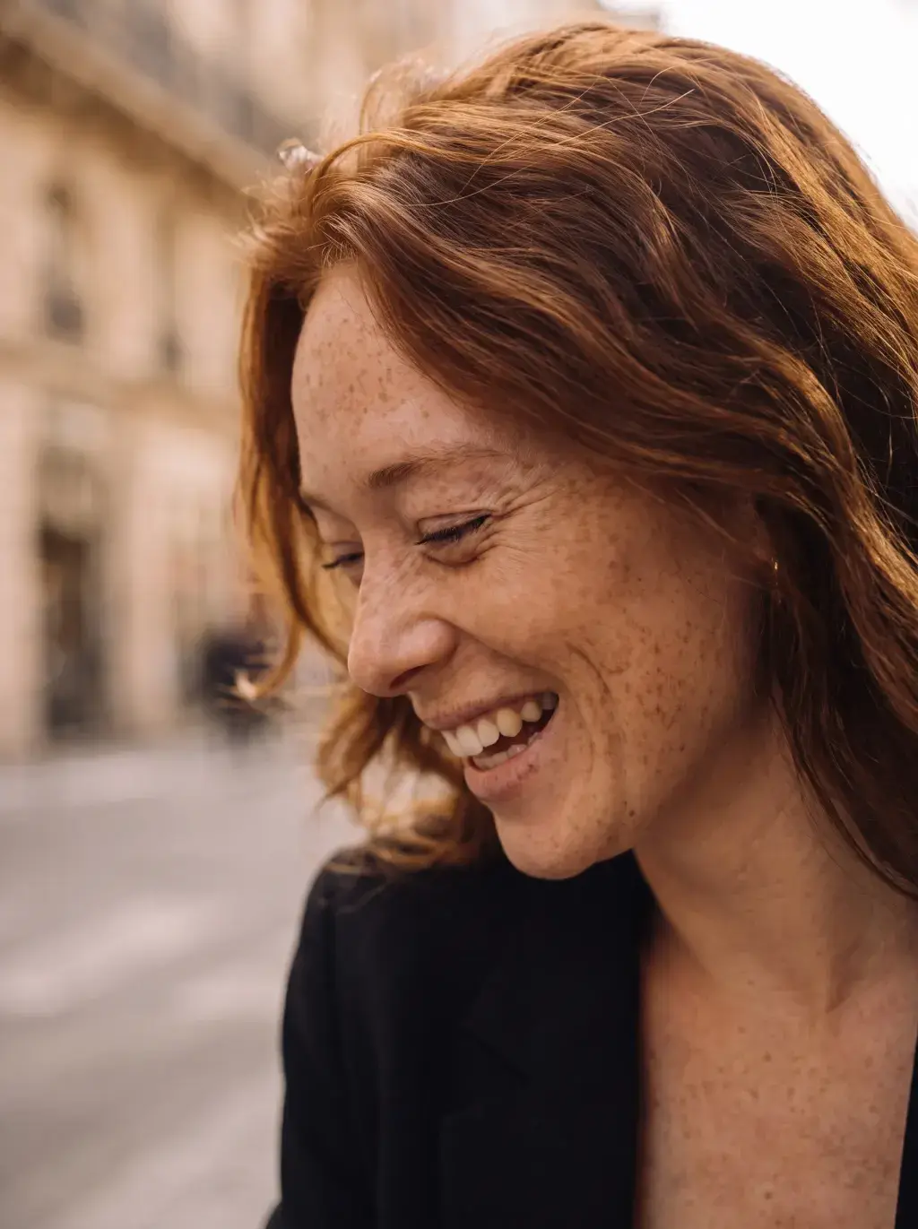 Portrait de profil d'une femme rousse souriante en blazer noir dans une rue parisienne