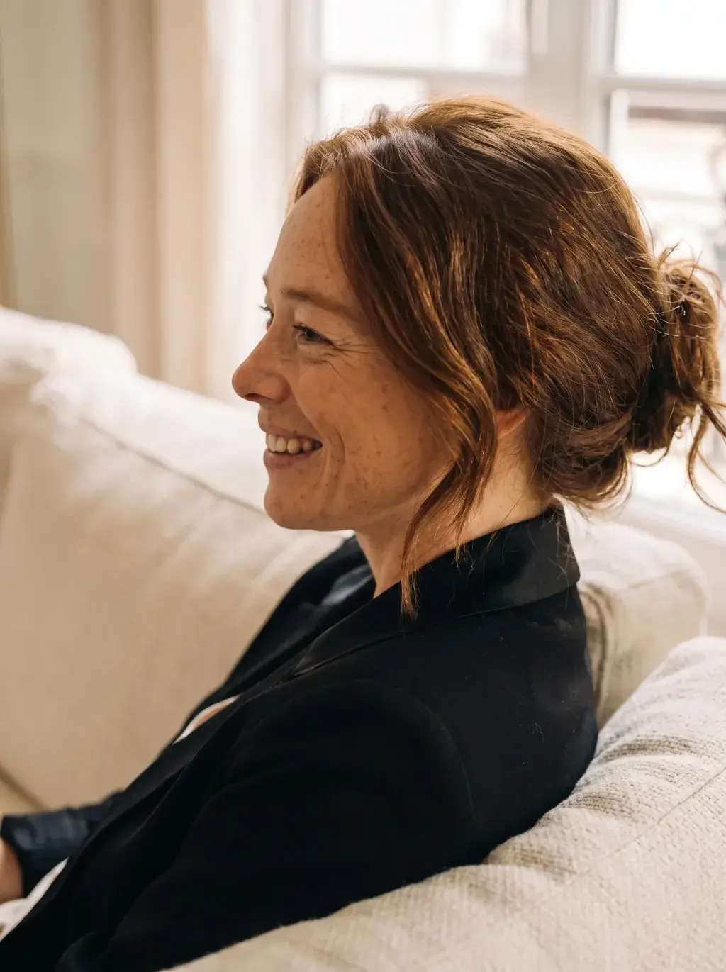 Portrait de profil d'une femme rousse souriante en blazer noir sur un canapé crème