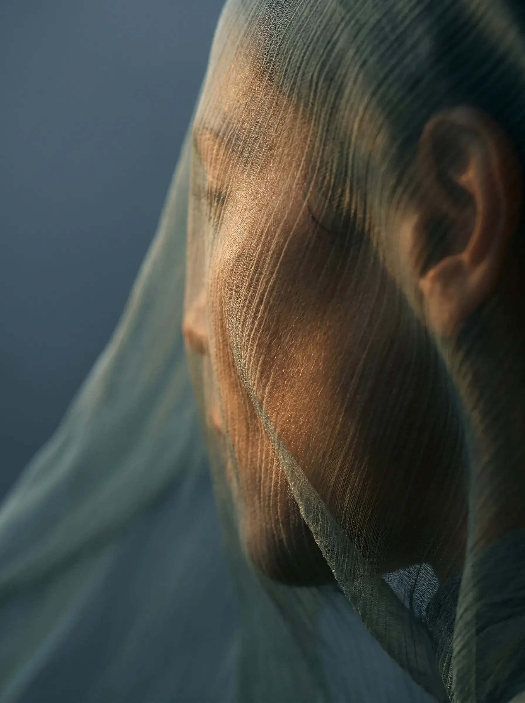 Portrait gros plan femme à travers voile gaze – lumière chaude dorée sur le visage, fond bleu-gris