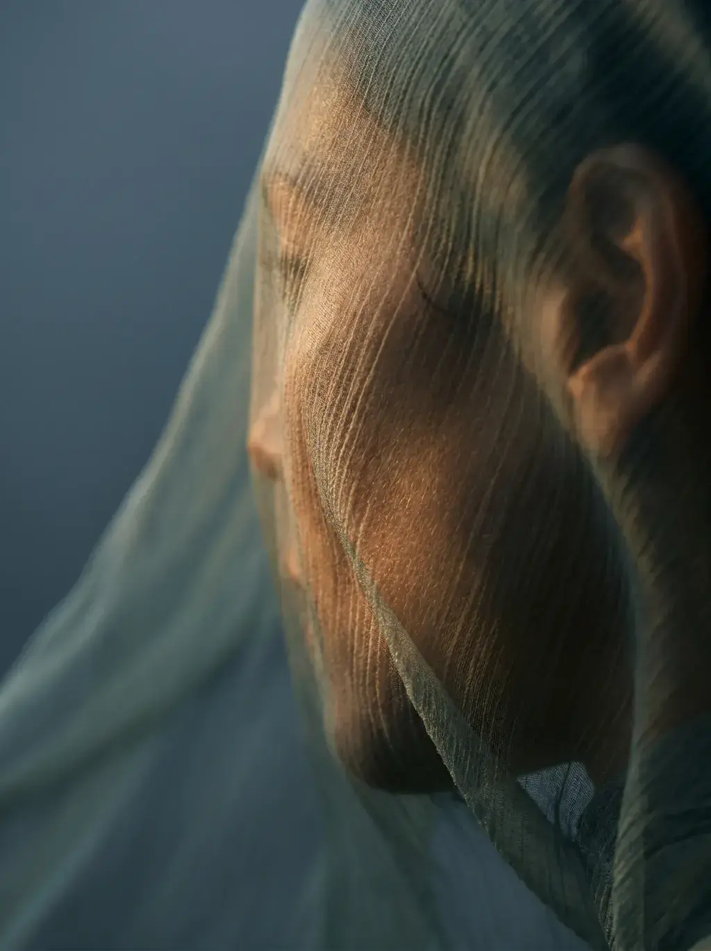 Portrait gros plan femme à travers voile gaze – lumière chaude dorée sur le visage, fond bleu-gris