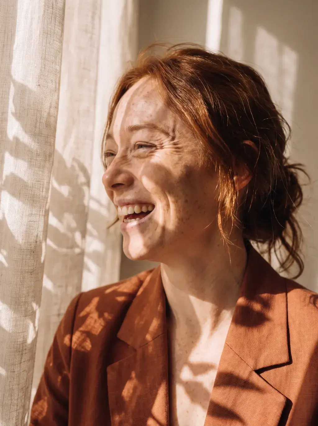 Portrait lumineux d'une femme rousse souriante en blazer terracotta avec jeu d'ombres de feuilles