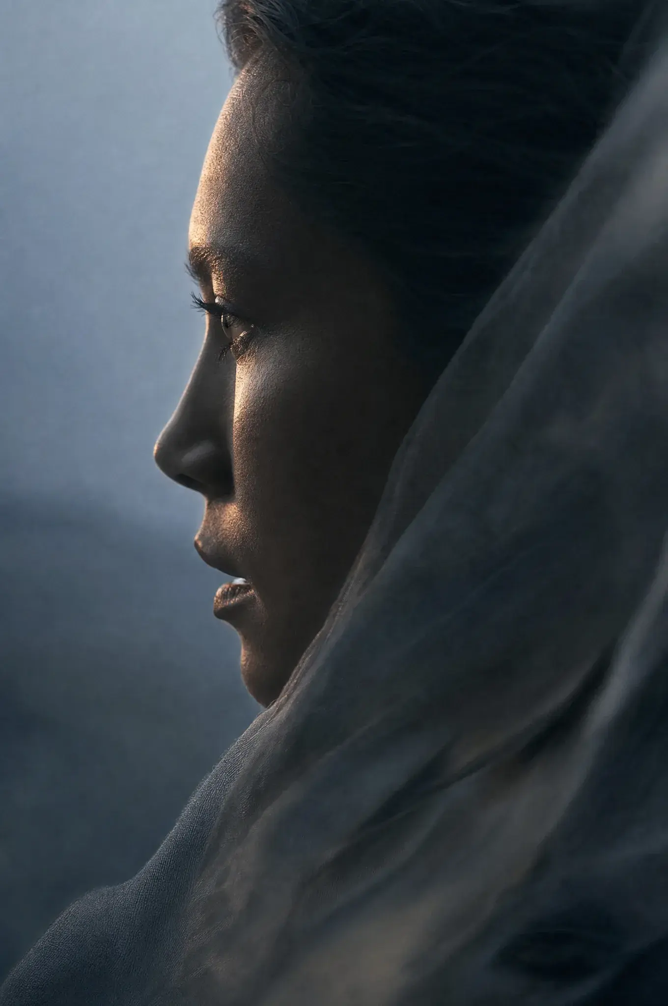 Portrait profil femme – voile gaze enveloppant tête et épaules, lumière chaude sur peau, fond bleu