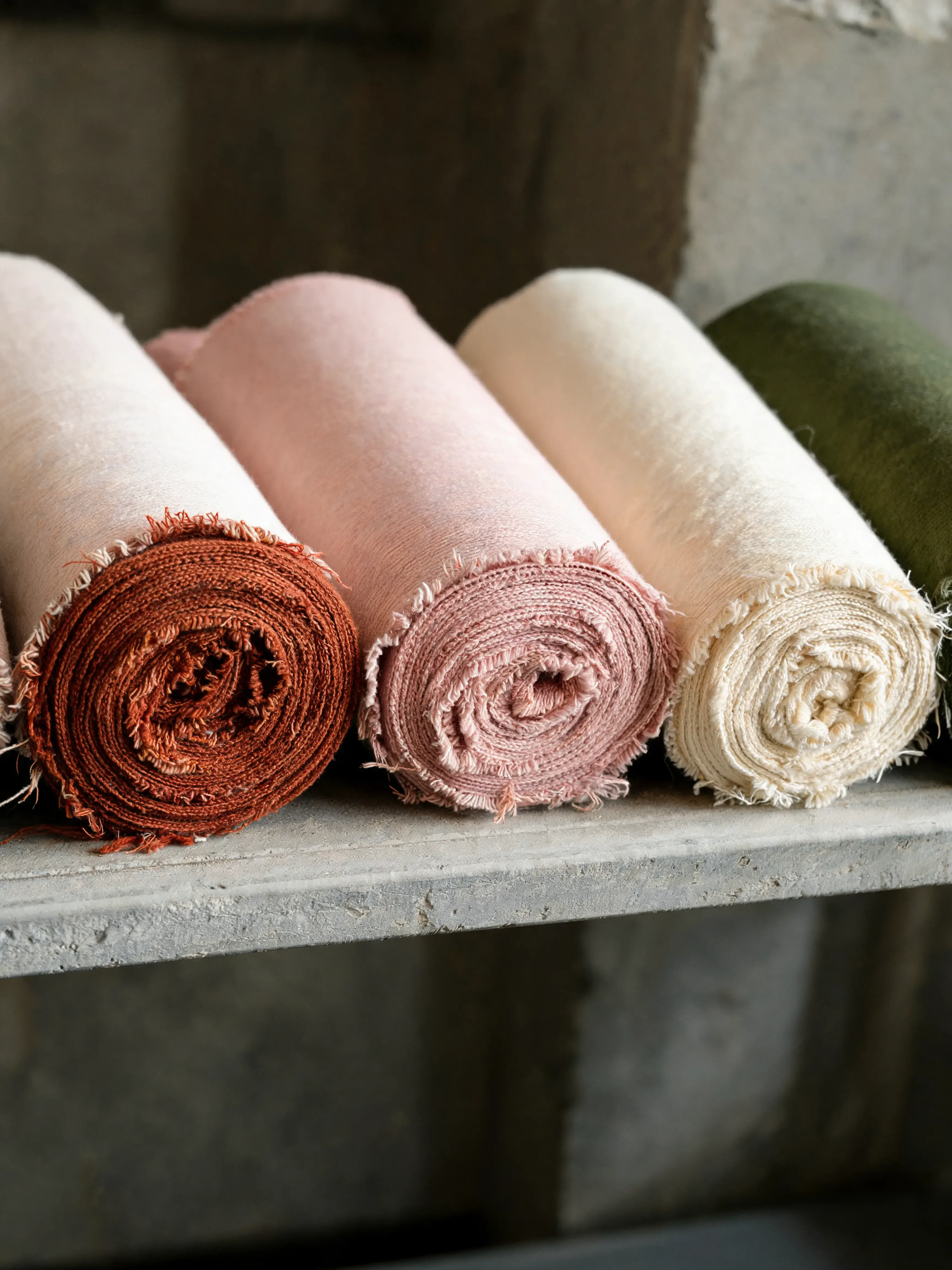 Quatre rouleaux tissu colorés sur étagère béton – terracotta rose crème vert, atelier textile