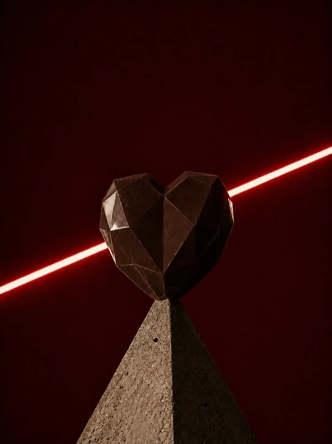 Sculpture de cœur géométrique facetté sombre sur socle béton avec rayon néon rouge traversant horizontalement, fond bordeaux
