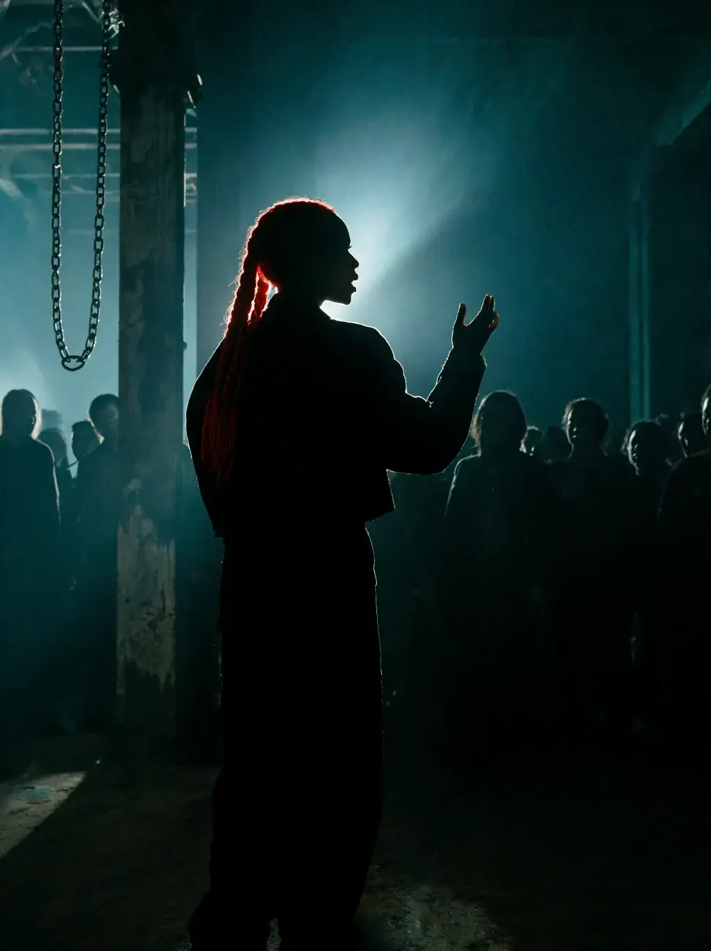 Silhouette de profil d'une femme aux tresses rouges prenant la parole devant un groupe dans un hangar