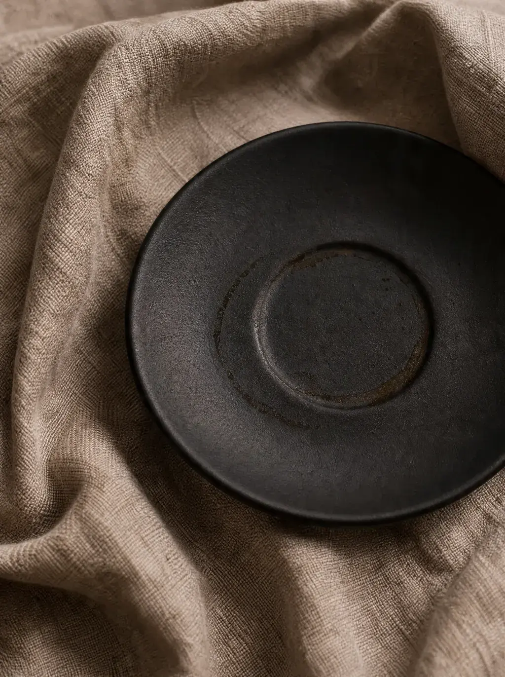 Sous-tasse céramique noire matte sur nappe lin froissée – composition minimaliste, fond beige