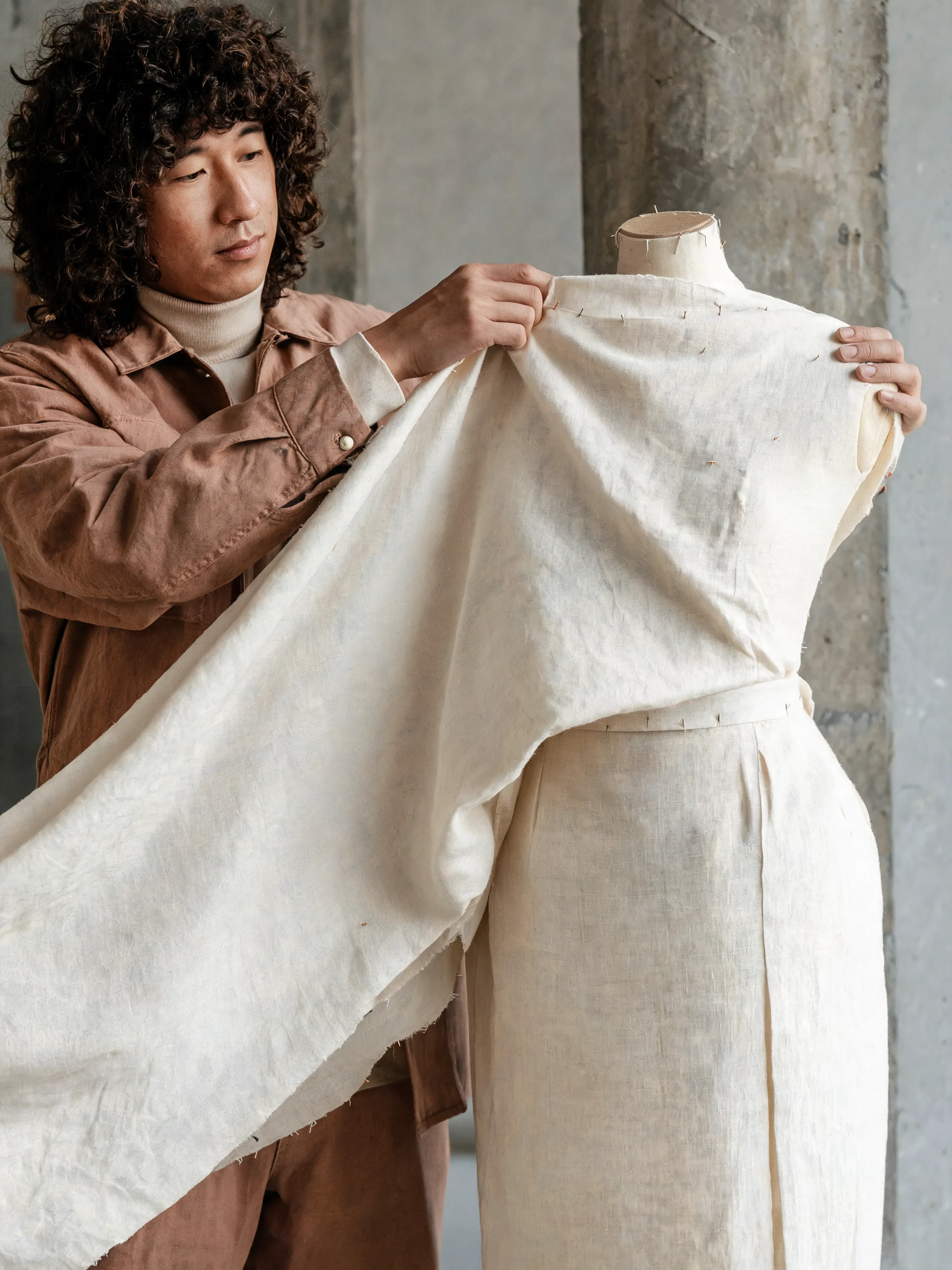 Styliste drapant tissu lin crème sur mannequin couture – pilier béton, mur studio, lumière naturelle
