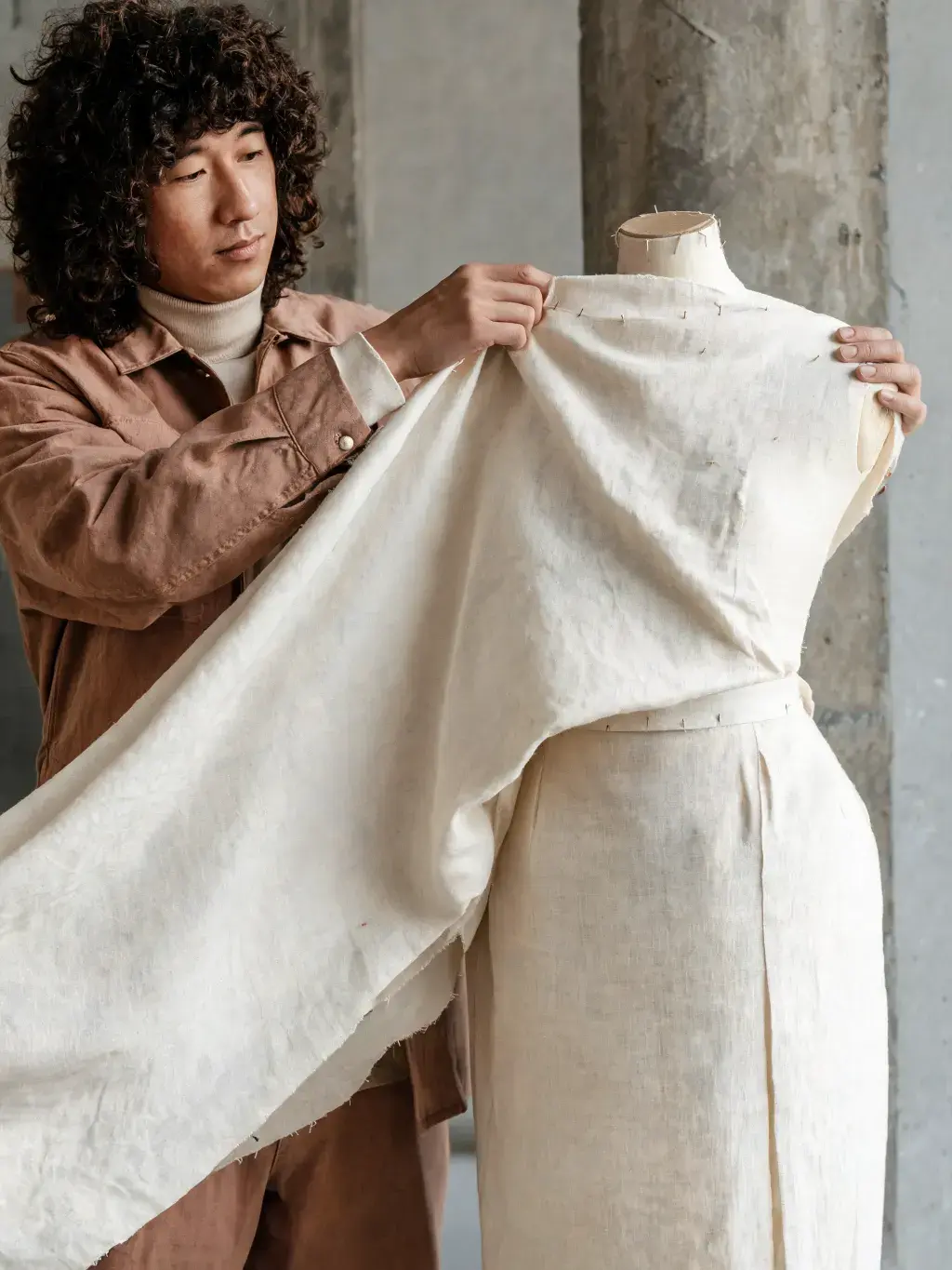 Styliste drapant tissu lin crème sur mannequin couture – pilier béton, mur studio, lumière naturelle
