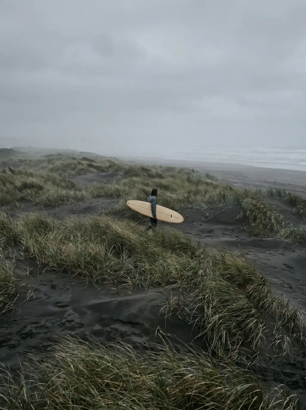 Surfeur portant une planche en bois marchant dans les dunes côtières sous ciel gris