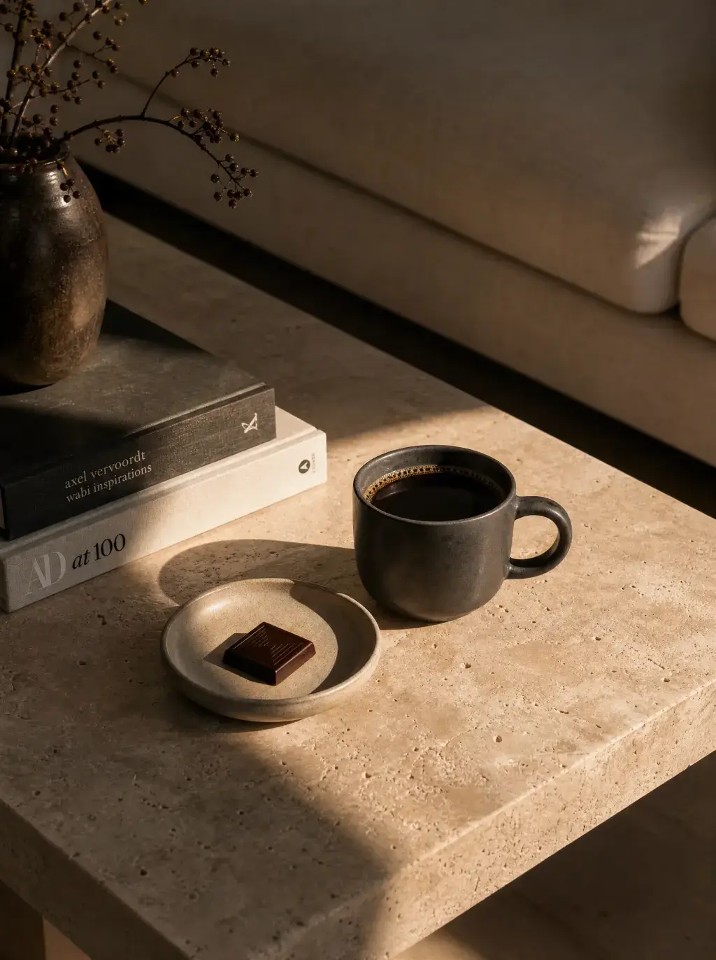 Table basse travertin – mug café noir, carré chocolat, livres d'art, vase soliflore, lumière rasante