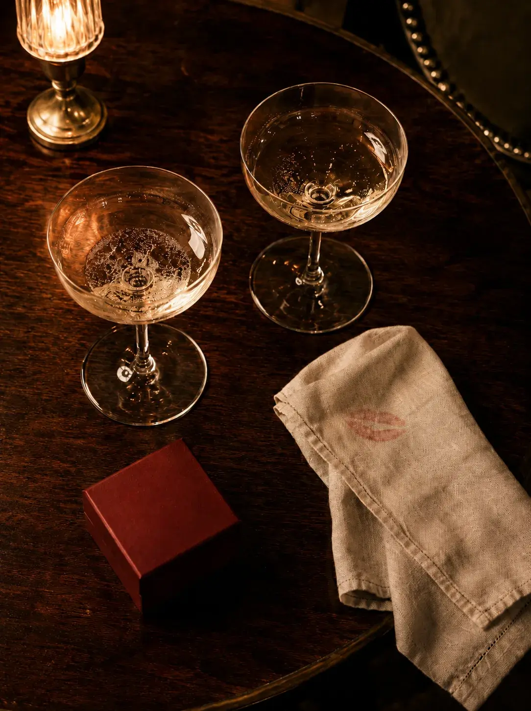 Table bistrot en bois foncé avec deux coupes de champagne, serviette lin avec empreinte de rouge à lèvres bordeaux, petite boîte rouge
