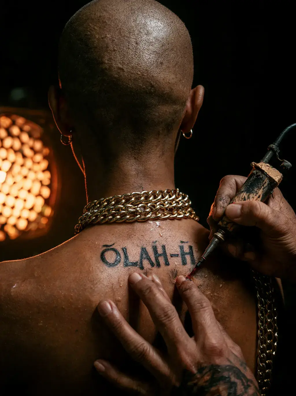 Tatoueur tatouing ÖLAH-H dans le dos d'un homme au crâne rasé portant une chaîne dorée