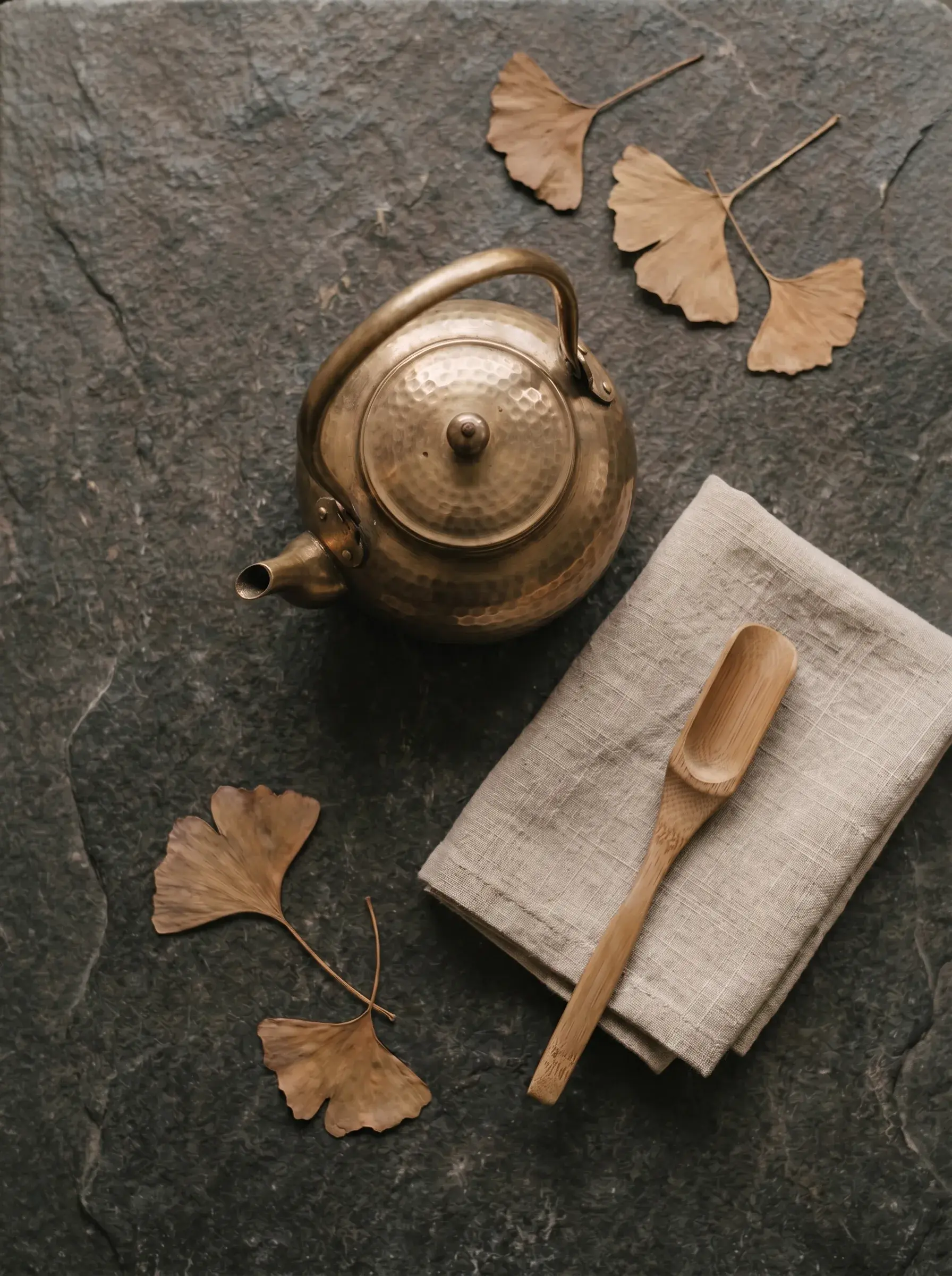Théière en bronze martelé sur ardoise noire avec feuilles de ginkgo séchées, linge lin et cuillère bambou