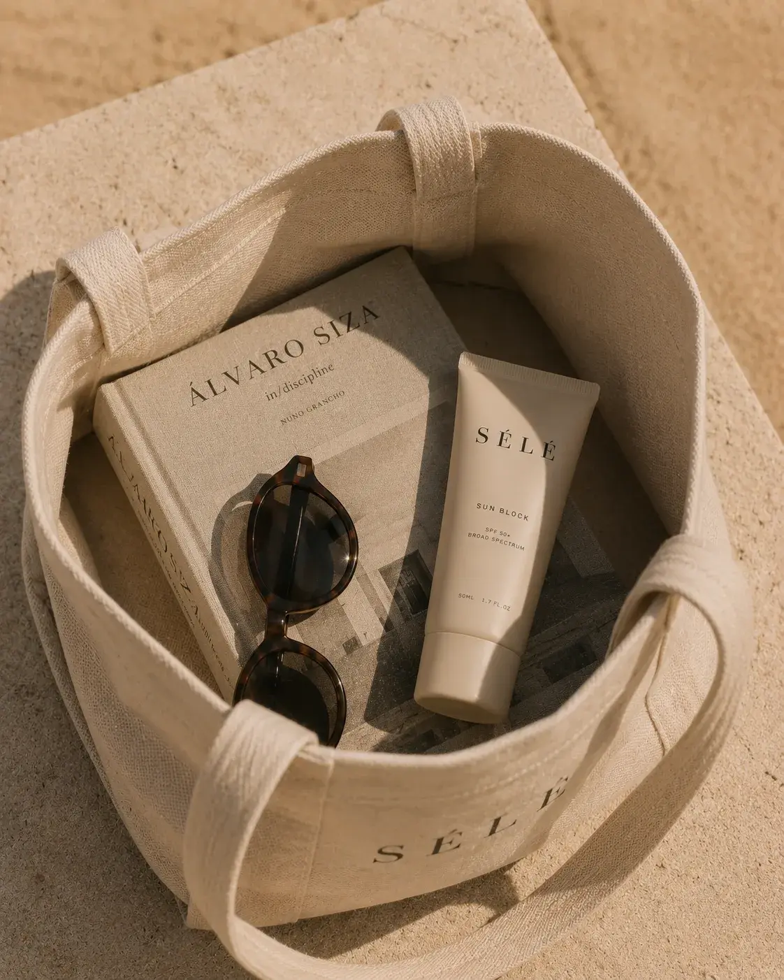 Tote bag blanc sur sable – tube crème solaire, lunettes de soleil, livre d'architecture, flatlay plage