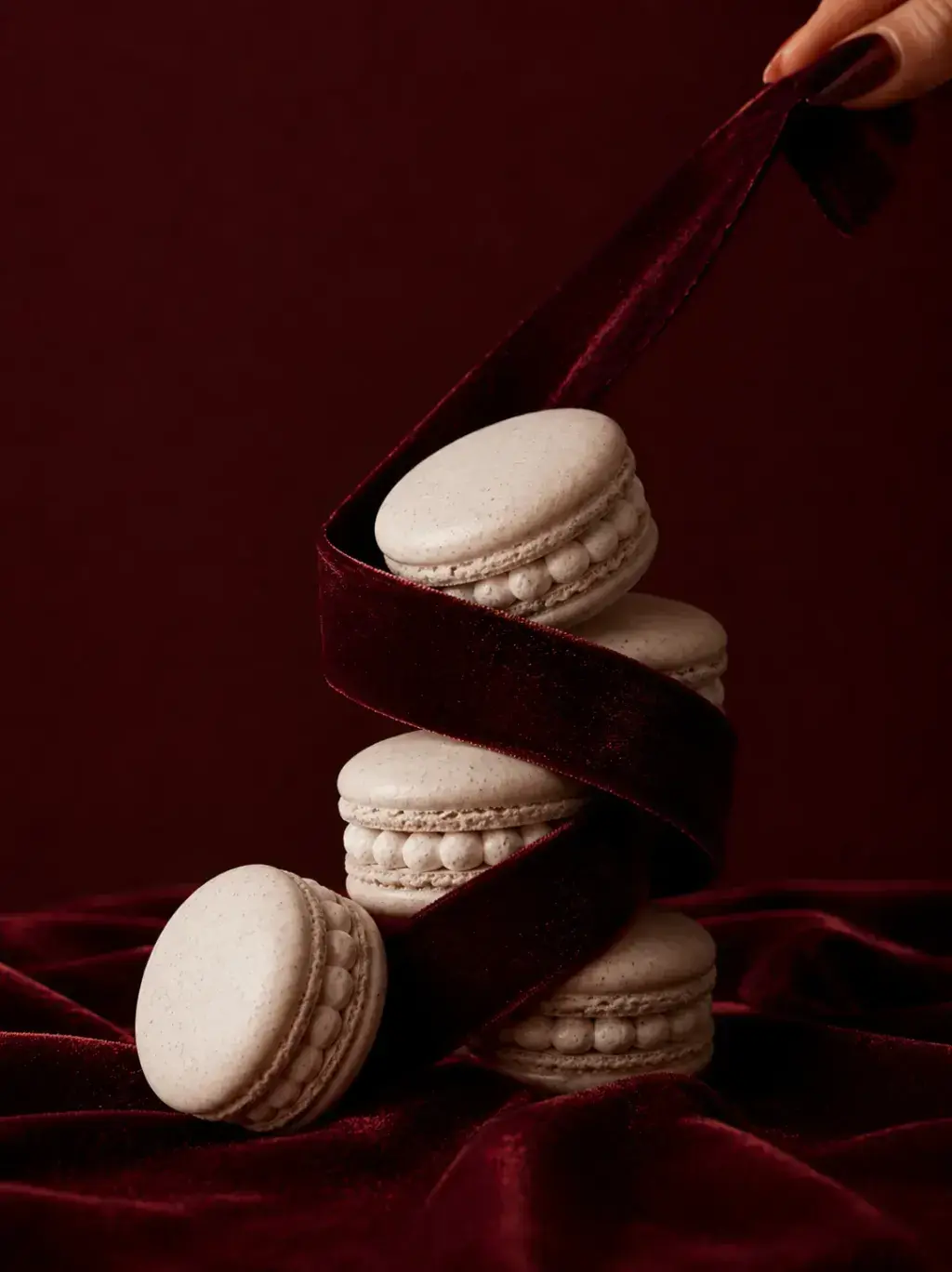 Tour de macarons blancs côtelés enlacés d'un ruban de velours bordeaux, main tirant le ruban, tissu bordeaux drapé en fond