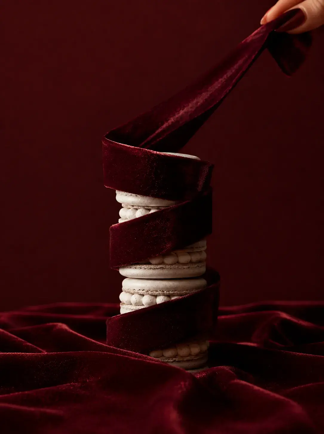 Tour de macarons blancs serrée enroulée d'un ruban de velours bordeaux déroulé vers le haut par une main, drapé bordeaux en fond