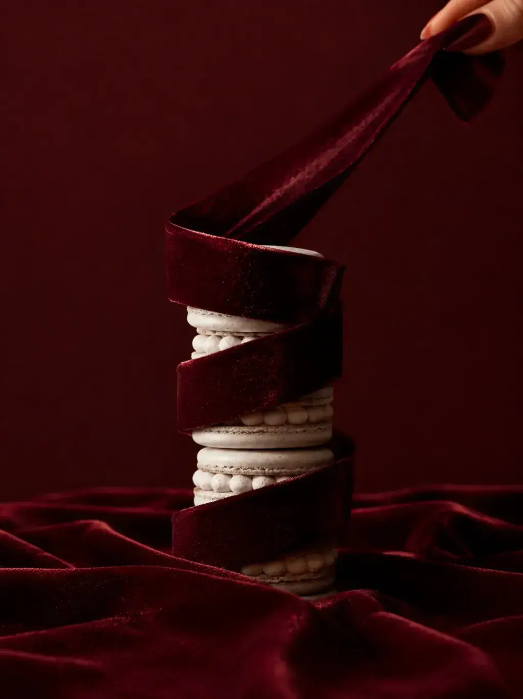 Tour de macarons blancs serrée enroulée d'un ruban de velours bordeaux déroulé vers le haut par une main, drapé bordeaux en fond