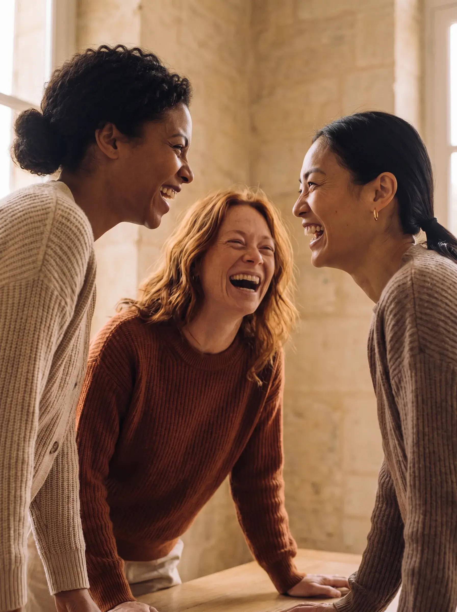 Trois femmes de trois origines riant ensemble autour d'une table dans un espace en pierre