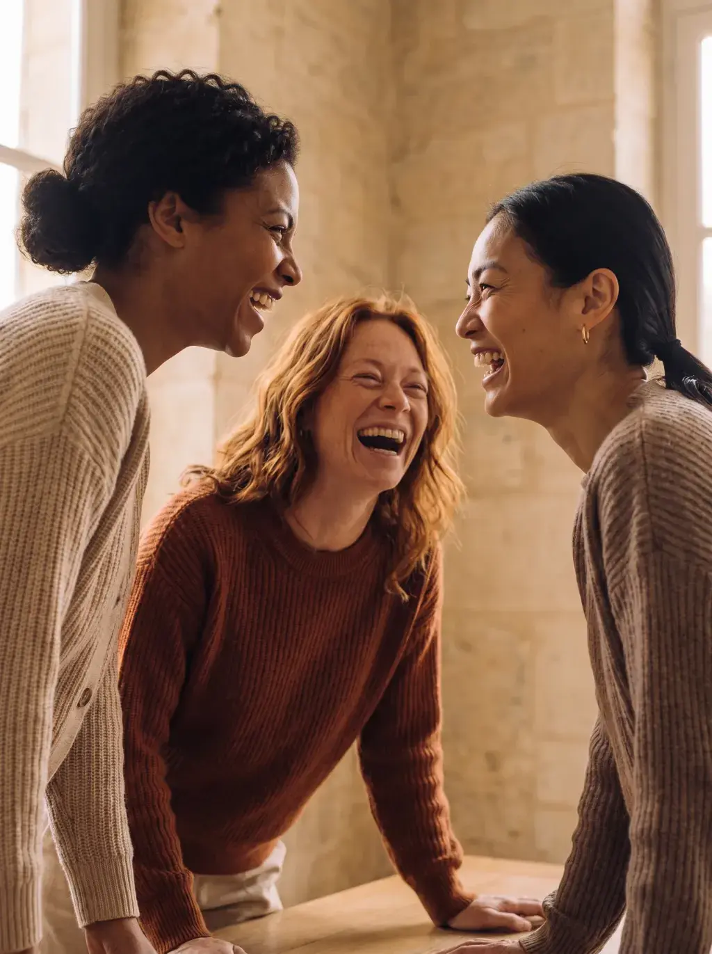Trois femmes de trois origines riant ensemble autour d'une table dans un espace en pierre
