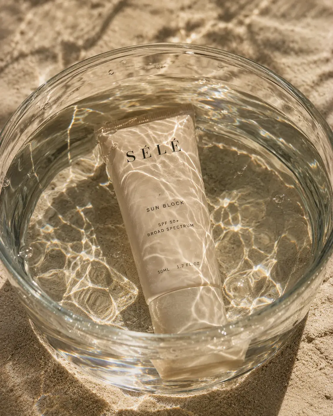 Tube crème solaire dans bol en verre d'eau – reflets aquatiques, sable, lumière zénithale