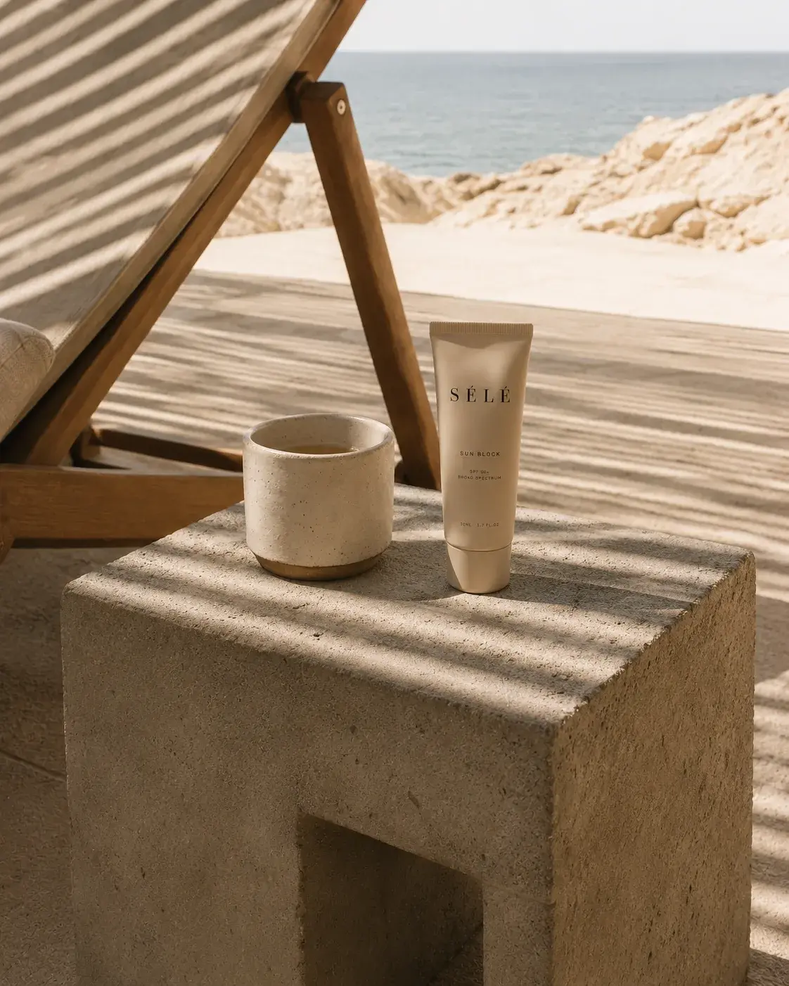 Tube crème solaire et mug céramique sur table basse béton – chaise longue, vue mer, ombres rayées