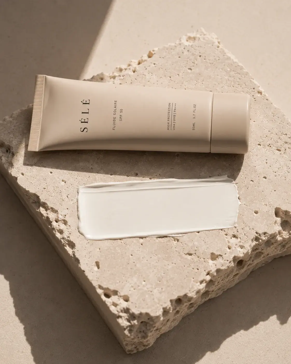 Tube crème solaire sur bloc travertin – trace de crème blanche, ombre végétale, fond neutre chaud