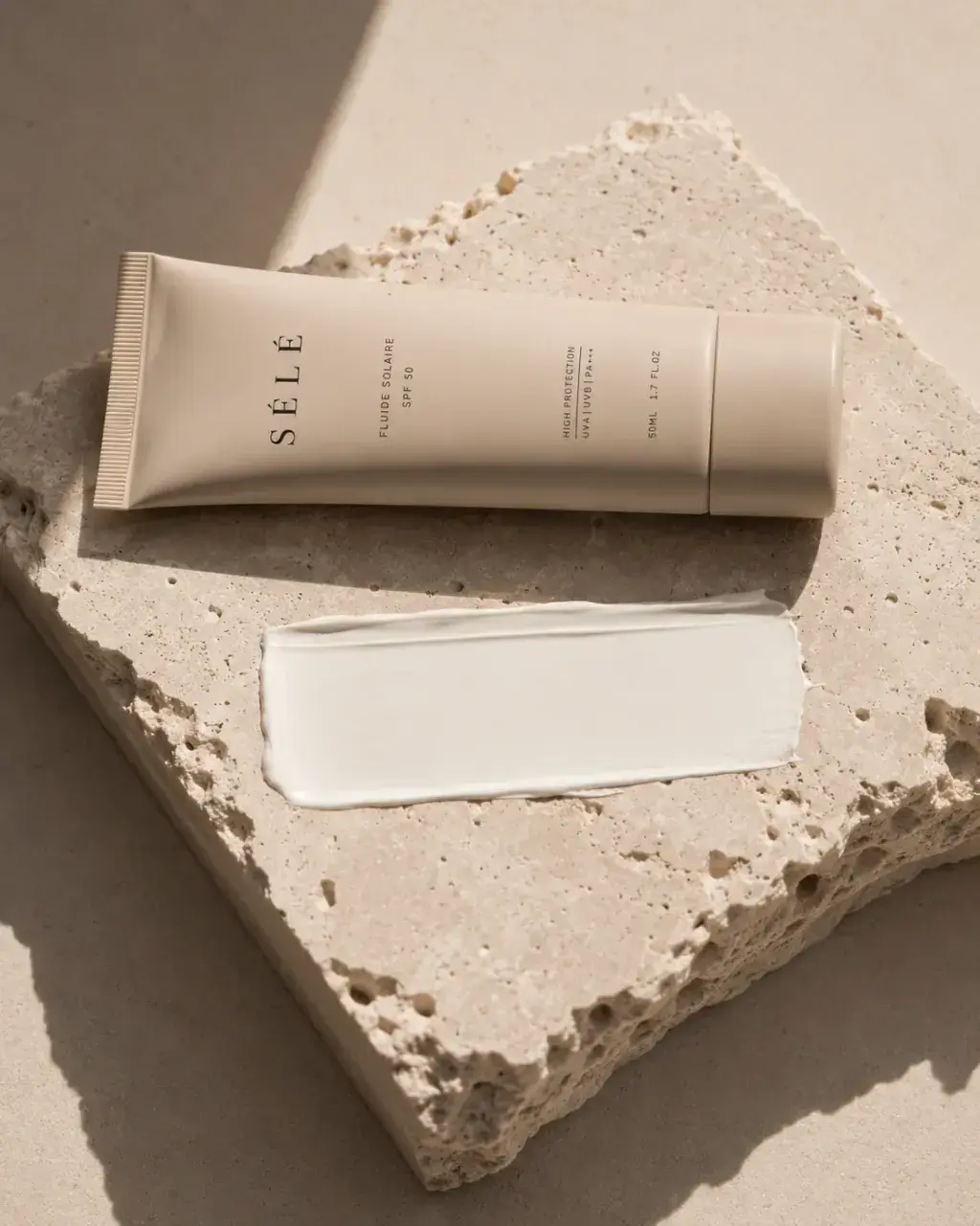 Tube crème solaire sur bloc travertin – trace de crème blanche, ombre végétale, fond neutre chaud