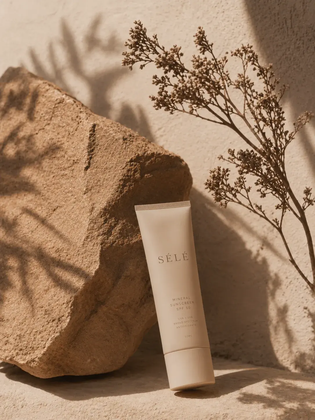 Tube crème SPF beige sur pierre – branche séchée, ombre végétale, mur plâtre sable