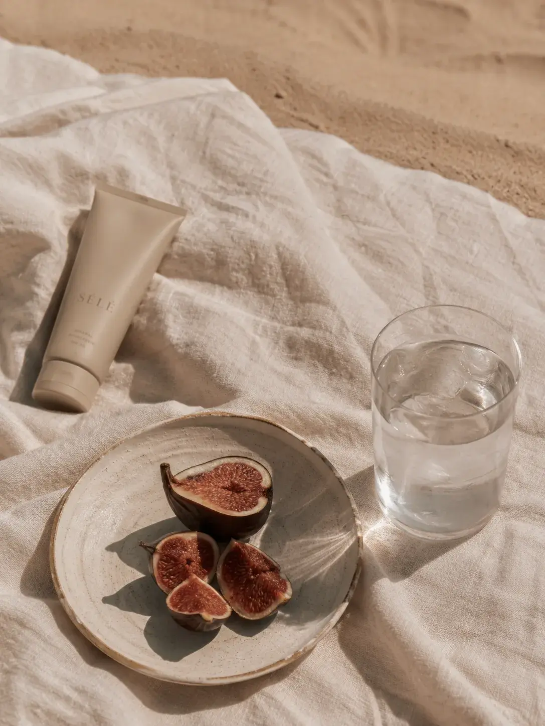 Tube de soin beige avec figues et verre d'eau sur linge de plage – sable, lumière naturelle