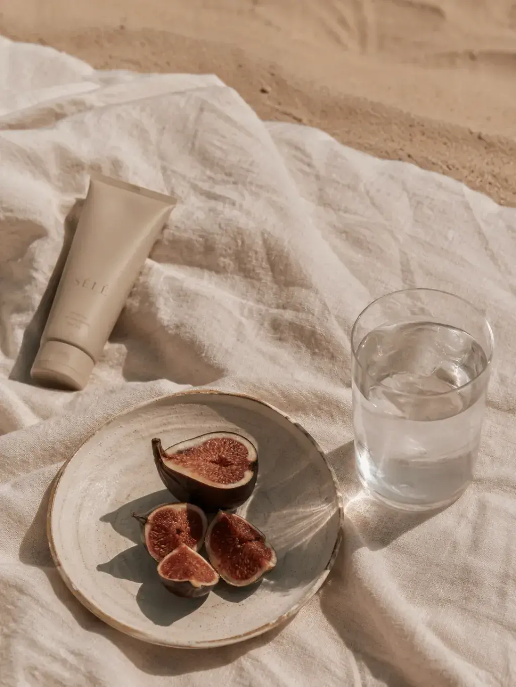Tube de soin beige avec figues et verre d'eau sur linge de plage – sable, lumière naturelle