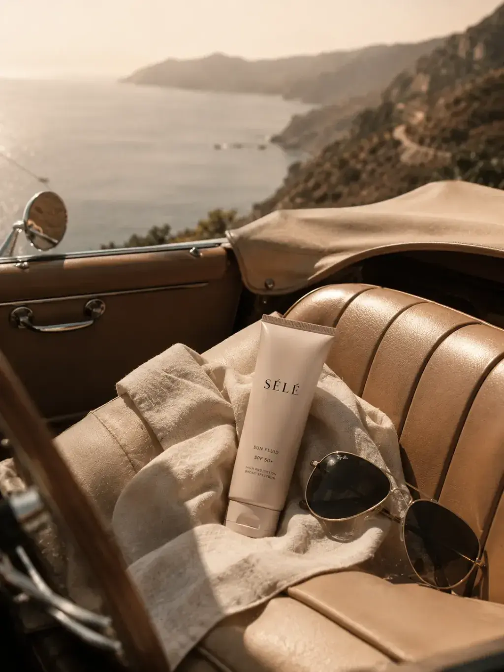 Tube fluide solaire sur siège cuir caramel de décapotable – serviette lin, lunettes, vue mer et collines