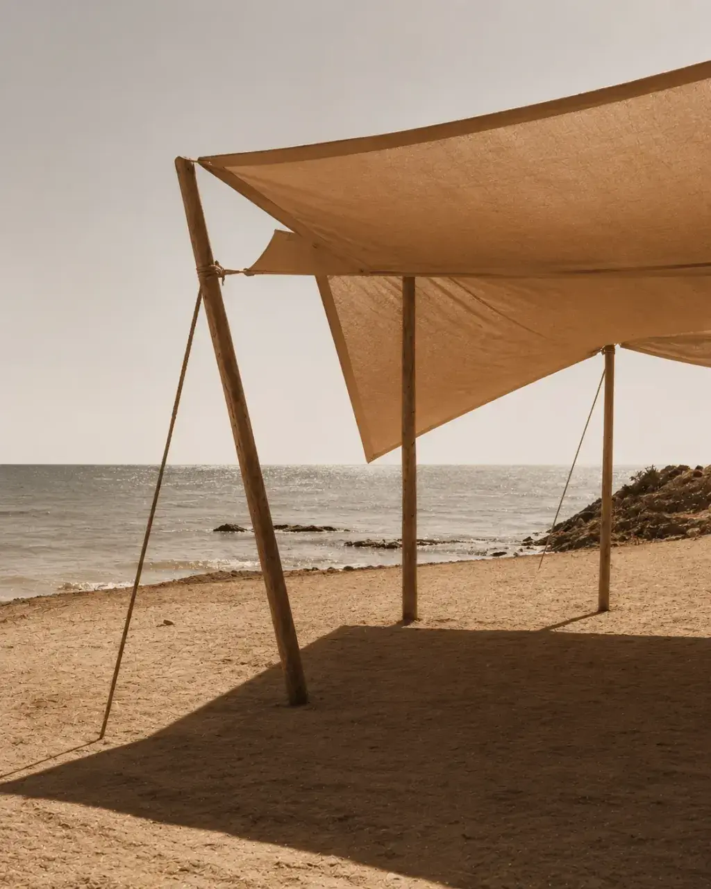 Voile de lin beige sur poteaux bambou en bord de plage – mer, rochers, ciel terne, ombre portée