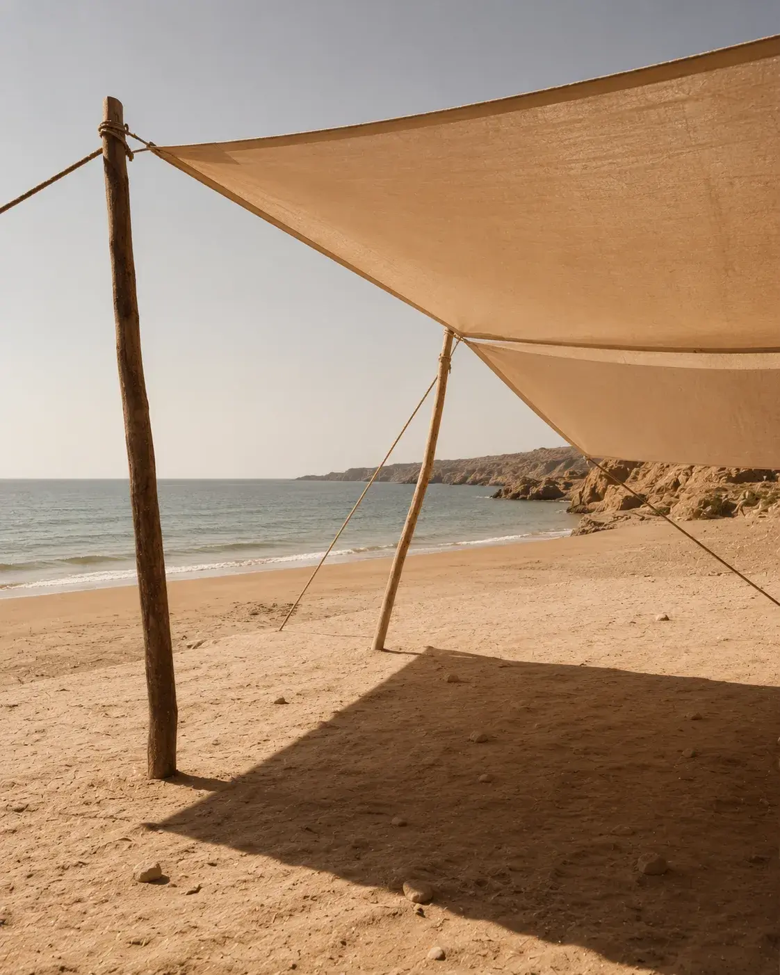 Voile en lin beige tendue sur poteaux en bois sur plage désertique – mer, falaises, ombre