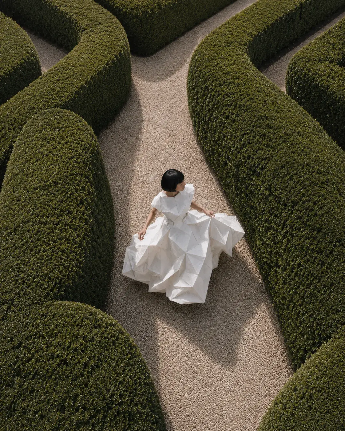 Vue aérienne femme en robe origami dans labyrinthe de haies courbes – ombre portée sur gravier