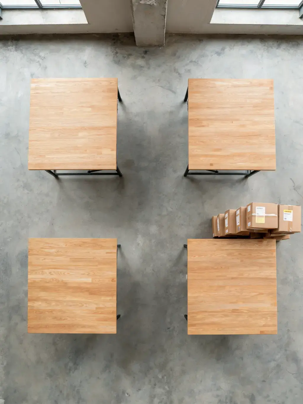 Vue aérienne quatre tables bois carrées – sol béton, cartons empilés, lumière zénithale, entrepôt