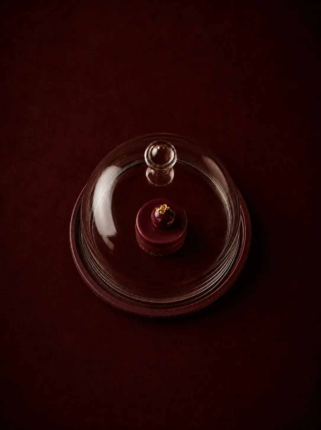 Vue aérienne sur petite pâtisserie rouge individuelle sous cloche en verre transparente, fond bordeaux profond, composition minimaliste