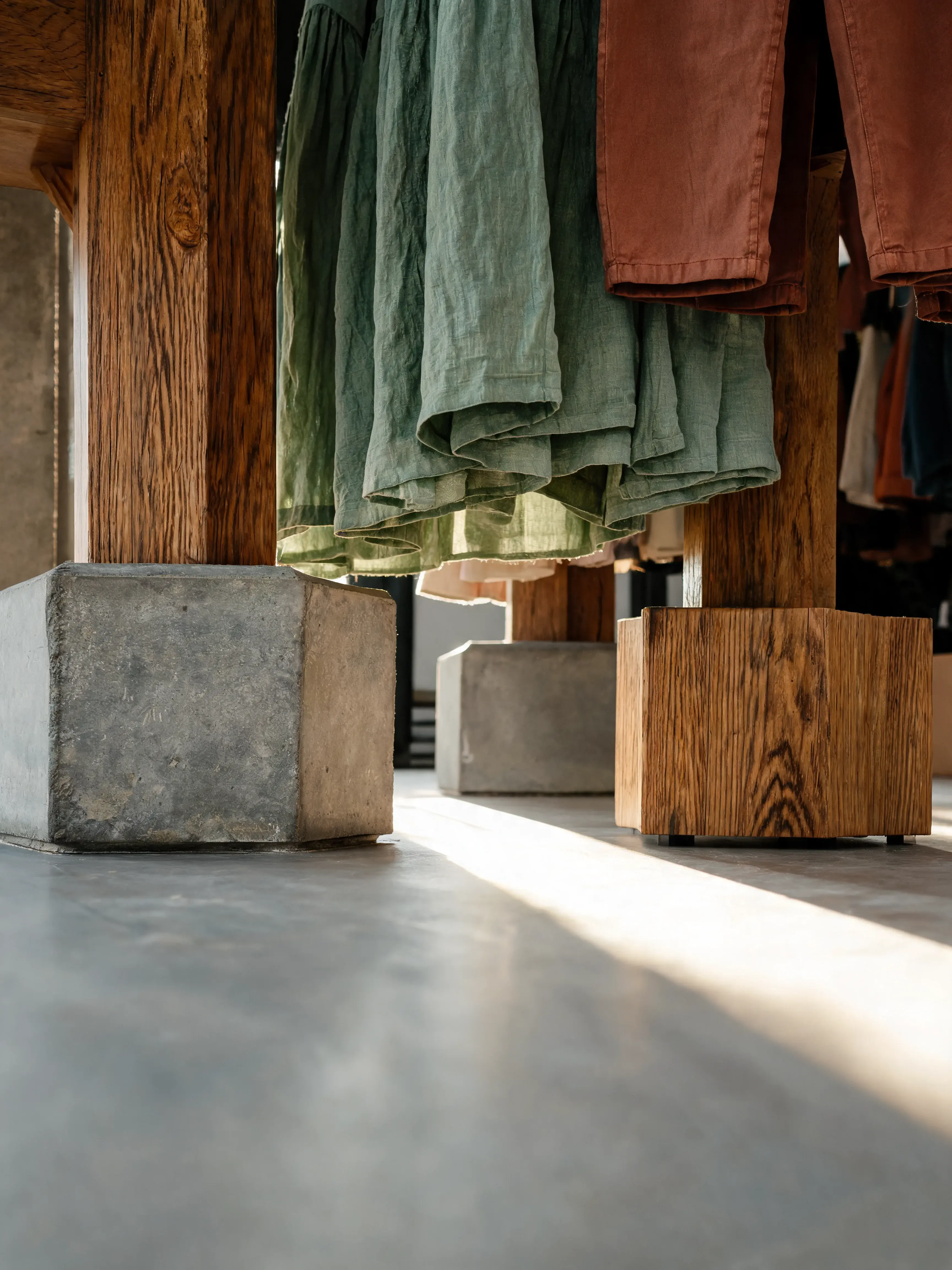 Vue basse portants mode – vêtements vert terracotta, socles béton bois, rayons lumière sol