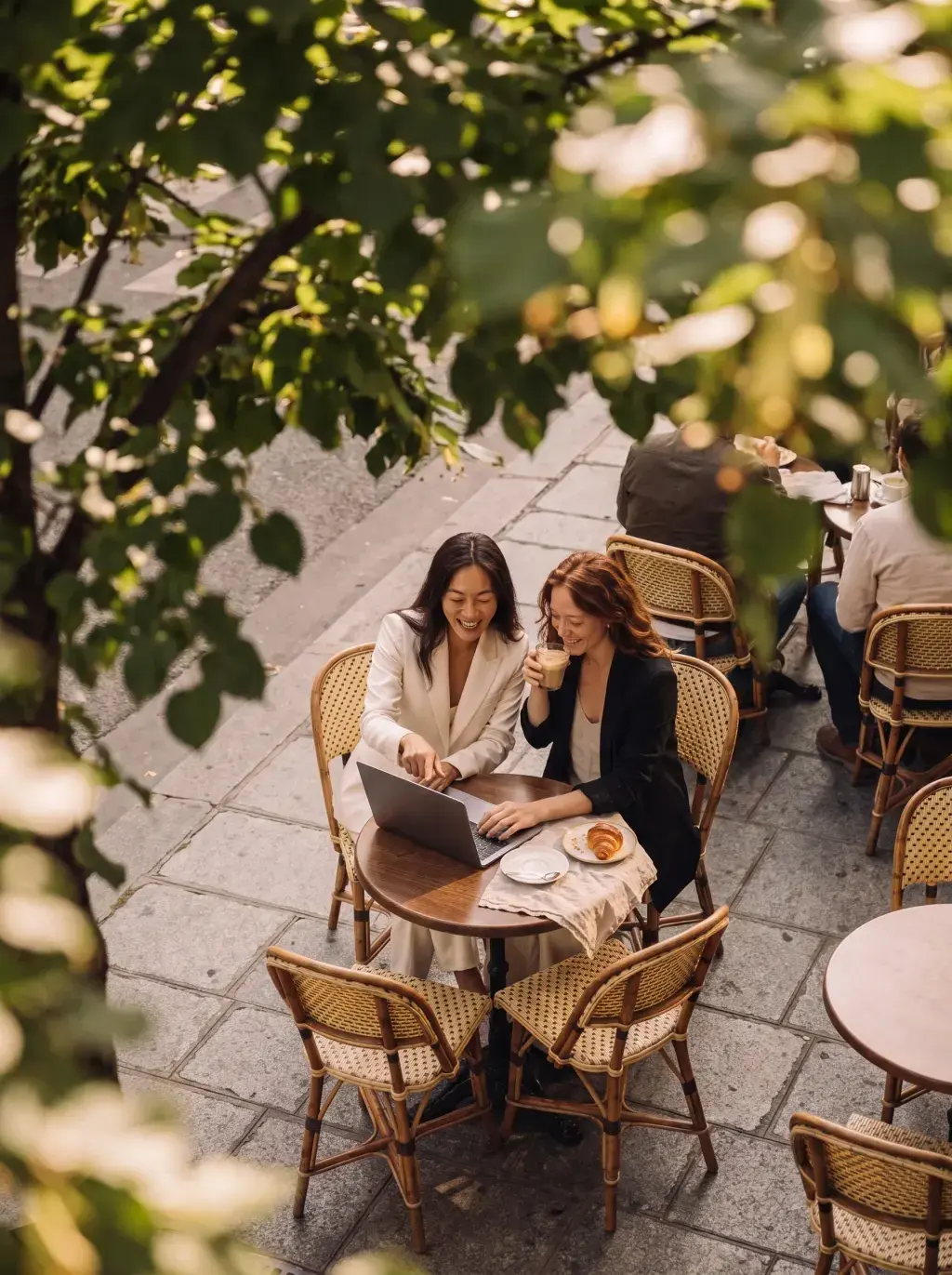 Vue en plongée de deux femmes consultant un laptop en terrasse de café encadrée de verdure