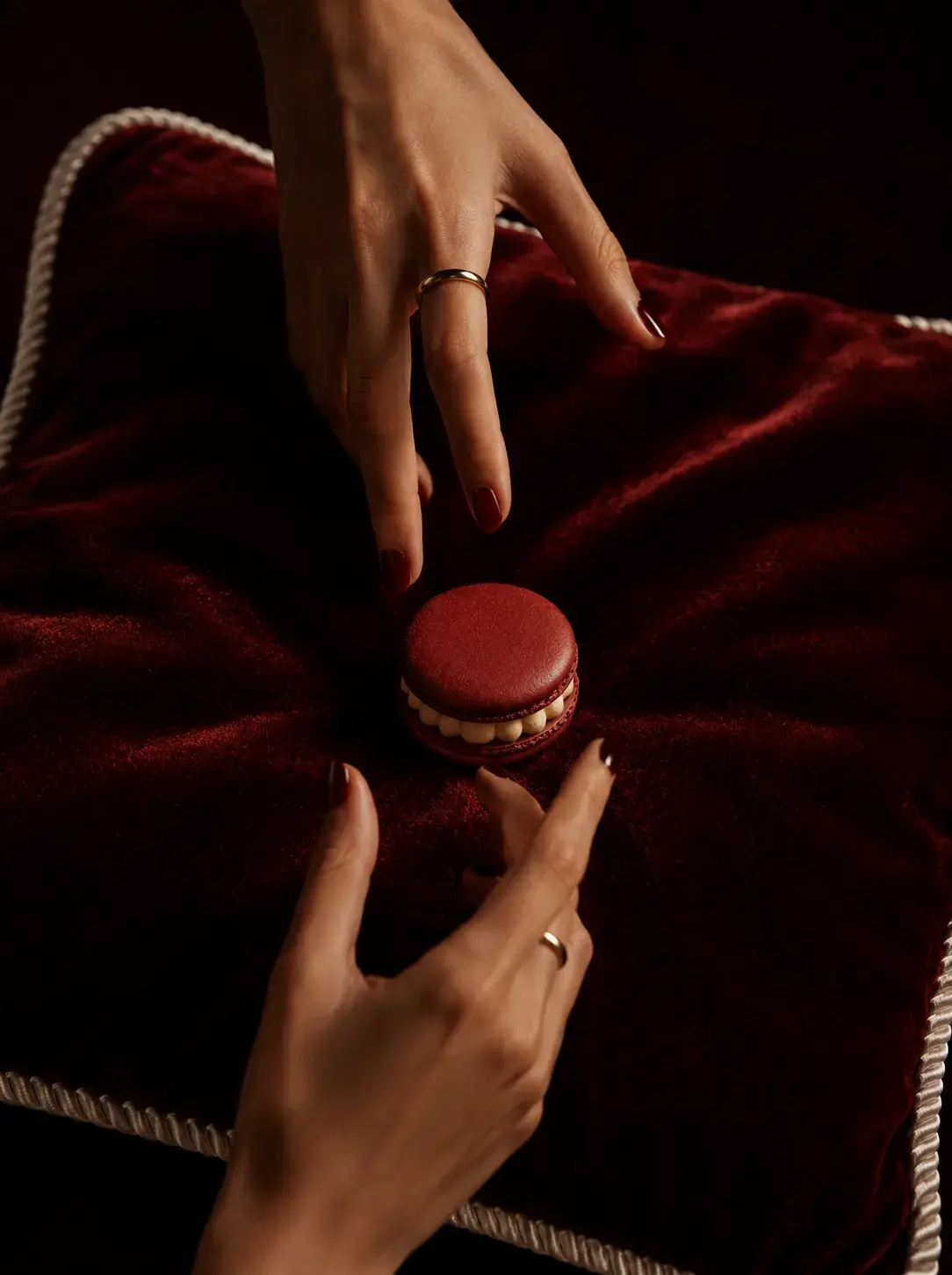 Vue plongeante sur deux mains aux bagues dorées saisissant un macaron bordeaux sur coussin de velours bordeaux