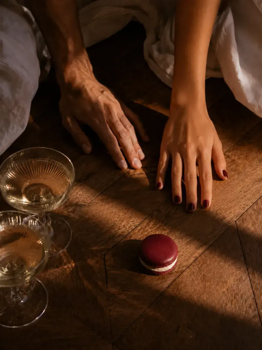 Vue plongeante sur deux paires de mains proches posées sur parquet avec verres de champagne couchés et macaron bordeaux