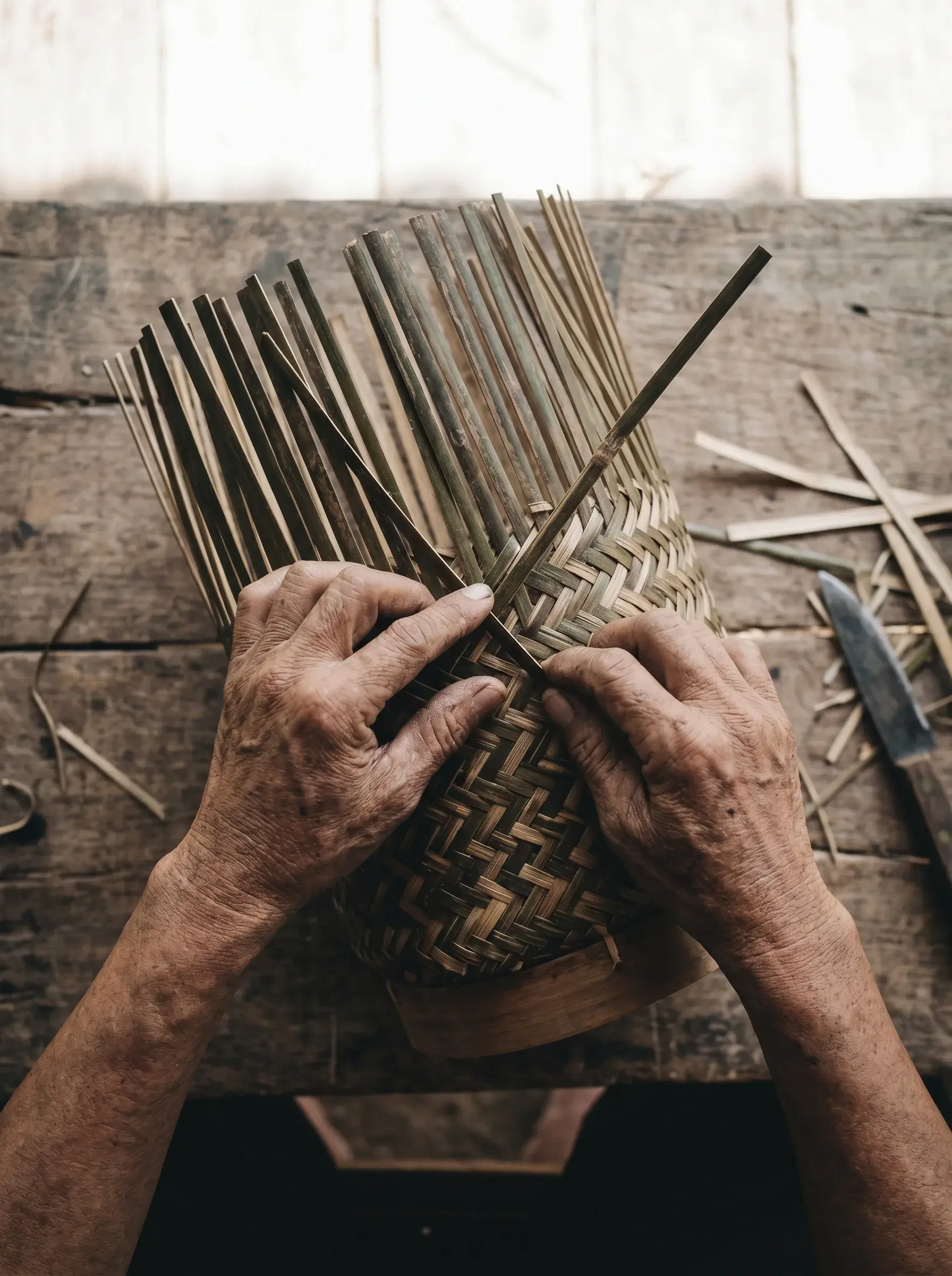 Vue plongeante sur mains ridées d'artisan tressant un panier en bambou sur table bois, lattes de bambou éparpillées