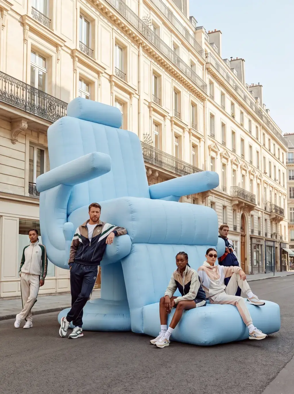 Groupe en survêtements pastel posant autour d'un fauteuil de barbier gonflable géant, rue parisienne
